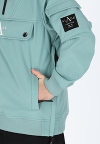 Helles türkisfarbiges Softshell-Jacke mit gerippten Bündchen und Saum, ausgestattet mit einer Chest-Tasche und einem schwarzen Logo-Patch am Oberarm.