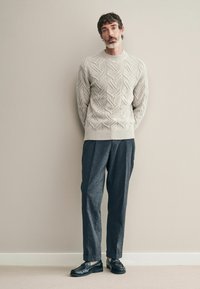 Hellgrauer kabelgestrickter Pullover, dunkelgraue Hose und schwarze Slipper. Neutraler Hintergrund. Modell steht mit hängenden Armen.