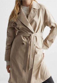 Manteau long beige avec un large col, boutons double-breasted et une ceinture à la taille cintrée. Tissu lisse avec un léger brillant.