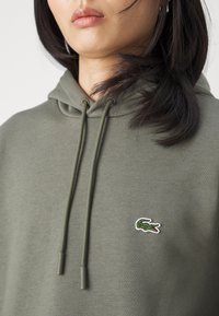 Donna che indossa una felpa con cappuccio verde oliva con il logo del coccodrillo Lacoste sul petto, primo piano che mostra i lacci della felpa e parte del volto.