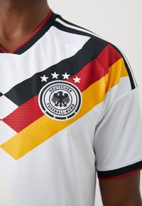 Nahaufnahme einer Person, die ein weißes Deutschland-Fußballtrikot mit schwarzen, roten und gelben diagonalen Streifen und dem Logo des Deutschen Fußball-Bundes trägt.