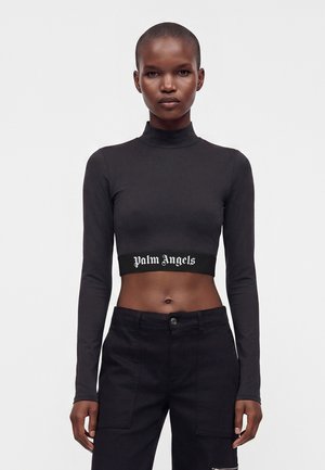 Γυναίκα που φοράει μαύρο crop top με μακριά μανίκια με κείμενο "Palm Angels" και μαύρο ψηλόμεσο παντελόνι στέκεται μπροστά σε λευκό φόντο.