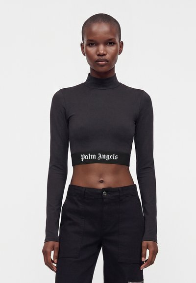 Γυναίκα που φοράει μαύρο crop top με μακριά μανίκια με κείμενο "Palm Angels" και μαύρο ψηλόμεσο παντελόνι στέκεται μπροστά σε λευκό φόντο.