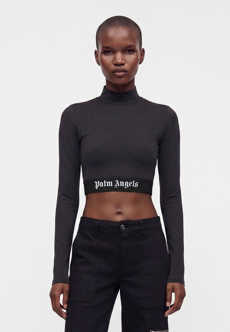 Γυναίκα που φοράει μαύρο crop top με μακριά μανίκια με κείμενο "Palm Angels" και μαύρο ψηλόμεσο παντελόνι στέκεται μπροστά σε λευκό φόντο.