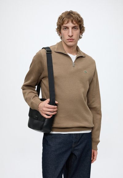 Jeune homme portant un pull zippé marron et un jean foncé tenant un sac à épaule noir sur un fond blanc uni.