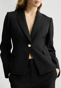 Blazer nero realizzato in tessuto liscio, con scollatura profonda, chiusura anteriore con bottoni, due tasche e revers affilati.