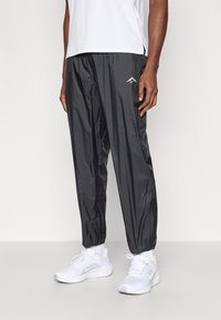 Μαύρο αδιάβροχο παντελόνι από ελαφρύ, γυαλιστερό υλικό με κωνικά πόδια και ελαστικές μανσέτες, συνδυασμένο με λευκά αθλητικά παπούτσια.