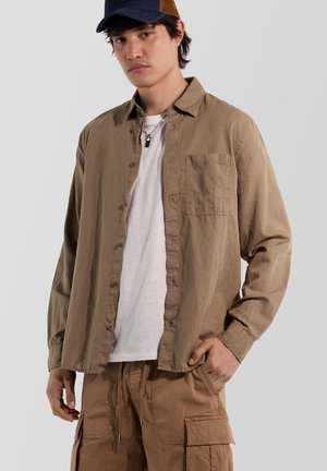 Junger Mann mit einer dunkelblauen Kappe, locker sitzendem hellbraunem Hemd über einem weißen T-Shirt und hellbraunen Cargohosen, steht mit einer Hand in der Hosentasche.