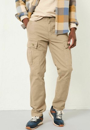 Beige Cargo-Hosen aus leichtem Material, mit Seitentaschen, genähten Details und einer lockeren Passform. Getragen mit marineblauen und braunen Turnschuhen.