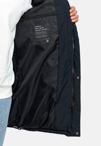 Donker marineblauwe jas met een gladde, duurzame buitenstof. De binnenkant heeft een glanzend zwarte voering en een zorglabel aan de linkerkant.