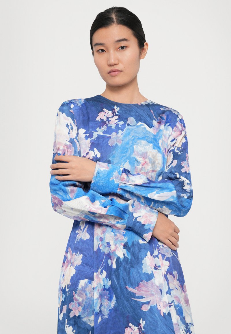 Vrouw met donker haar in een lange blauwe bloemetjesjurk, met gekruiste armen, staand tegen een effen lichte achtergrond.