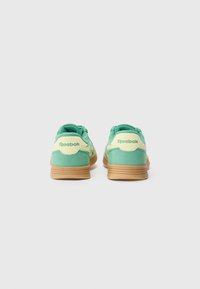 Sapatilhas verdes em camurça com sola e detalhes em bege. Apresentam uma forma arredondada, atacadores e o logótipo "Reebok" no calcanhar.