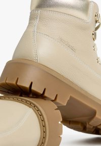 Bottes de randonnée en cuir beige avec un accent métallique doré, semelle rugueuse couleur tan et détails de couture. Logo imprimé sur le côté.
