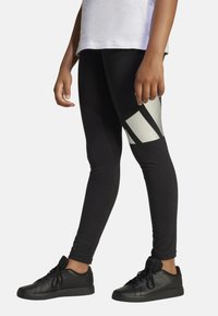 Leggings negros con acentos de patrón geométrico plateado. Hechos de material elástico, cuentan con un diseño ajustado. Combinados con zapatillas deportivas negras.