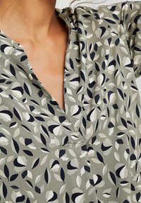 Gros plan sur une personne portant une robe avec des motifs de feuilles abstraits verts, blancs et bleu marine, et un décolleté en V.