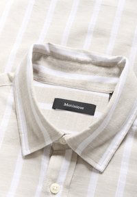 Col de chemise rayé beige et blanc à boutons avec une étiquette noire portant la mention Matinique à l’intérieur du col.