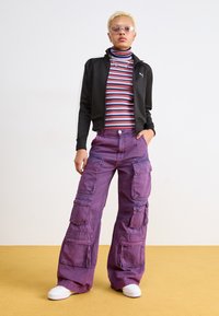 Black zip-up jacket with white accents, striped turtleneck, and purple cargo pants featuring multiple pockets. White sneakers complete the look.