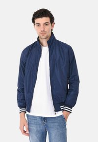 Giacca bomber blu navy con una texture liscia, colletto alto, zip completa e polsini e orlo a coste con righe. Indossata sopra una t-shirt bianca.