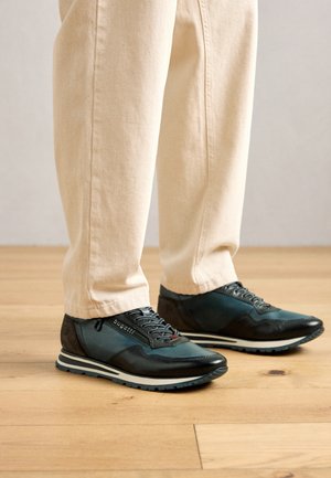 Sötétkék és fekete cipzáras fűzős sportcipőt viselő lábak bézs nadrággal, világos fa padlón állva.