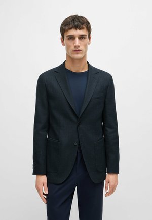 Blazer bleu foncé en chevron avec revers, fermeture à un bouton et deux poches avant, associé à une chemise bleu marine en dessous.