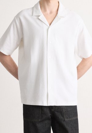 Chemise blanche à manches courtes en tissu texturé, dotée d'un col et de cinq boutons sur le devant ; portée avec un jean foncé.