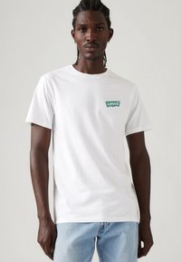 Mann med flettet hår iført en hvit Levi's t-skjorte og lyseblå jeans, som står mot en enkel lys grå bakgrunn.
