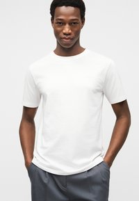 Λευκό βαμβακερό μπλουζάκι με στρογγυλή λαιμόκοψη και κοντά μανίκια, με διακριτικό λογότυπο στο στήθος, σε συνδυασμό με γκρι παντελόνια.