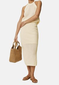 Strikket cremefarvet kjole med halterneck og tætsiddende silhouet, kombineret med en vævet tan-farvet håndtaske og lysebrune flade sandaler.
