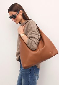 Brązowa skórzana torebka hobo o gładkiej fakturze i subtelnym logo, noszona swobodnie na beżowym swetrze i niebieskich dżinsach.