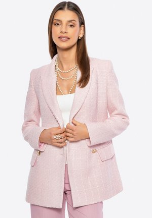 Donna che indossa un blazer testurizzato rosa chiaro, collane di perle sovrapposte, orecchini di perle, più anelli e pantaloni rosa su uno sfondo bianco.