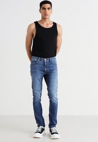 Crni majica bez rukava, plave slim-fit traperice i crno-bijele tenisice na muškom figuri protiv jednostavne bijele pozadine.