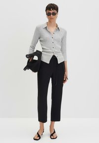 Camicetta grigia chiara con un motivo testurizzato, abbinata a pantaloni neri a sigaretta e sandali neri con punta aperta, mentre tiene una borsa nera.