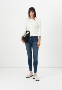 Haut blanc à manches longues avec des boutons, jean skinny bleu et chaussures slip-on crème. Tenant un sac à main noir, debout sur un fond uni.
