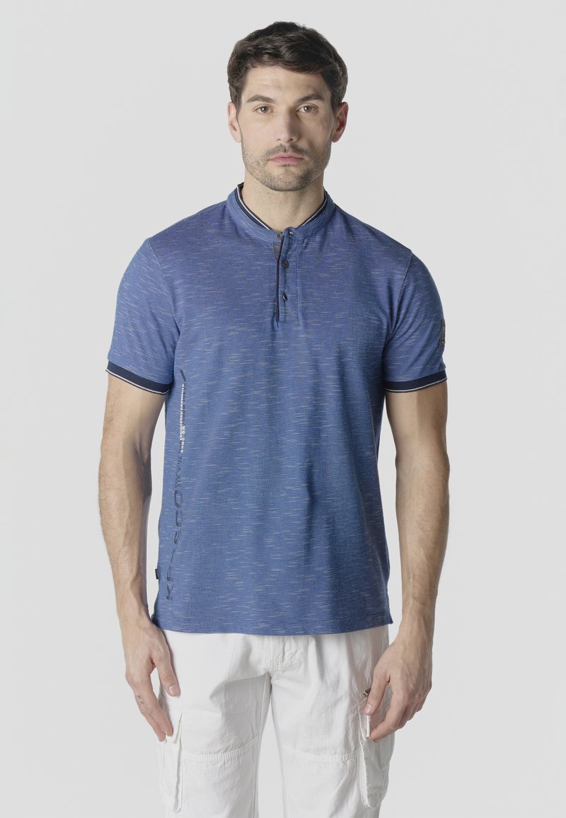 Homme portant un haut henley bleu à manches courtes avec patte de boutonnage et un pantalon cargo blanc, debout devant un fond uni.