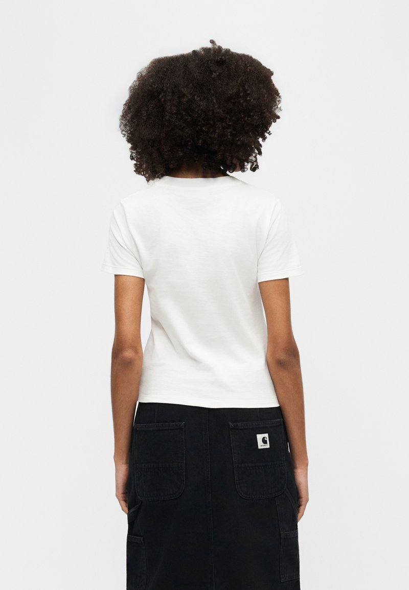 Persona con capelli ricci che indossa una maglietta bianca a maniche corte semplice e una gonna nera con grandi tasche posteriori, vista di spalle su uno sfondo bianco.