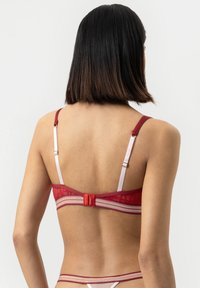 Vrouw met schouderlang donker haar in een rode bh en bijpassende string, gezien van achteren tegen een effen witte achtergrond.