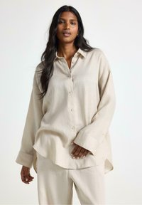 Lys beige, langærmet skjorte med knaplukning og afslappet pasform, lavet af et struktureret stof, med en buet kant og oversized manchetter.