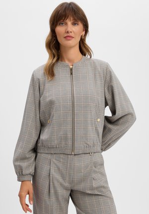Frau trägt eine grau karierte Reißverschlussjacke und passende Hose, steht mit einer Hand in der Hüfte vor einem schlichten weißen Hintergrund.