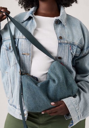 Sac bandoulière en denim à la forme douce et arrondie ; couleur teal profond ; bandoulière réglable ; fermeture éclair ; texture subtile. Porté sur un fond clair.