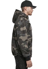 Chaqueta de camuflaje en tonos de verde y gris, con capucha, bolsillo con cremallera, puños elásticos y un corte holgado. El material parece suave.