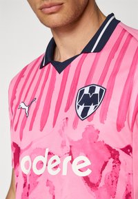 Rožinė sportinė marškinė su horizontaliais dryžiais ir abstrakčiomis raštais. Su tamsiai mėlynu apykakle, Puma logotipu ir ryškiu herbo ženkleliu.