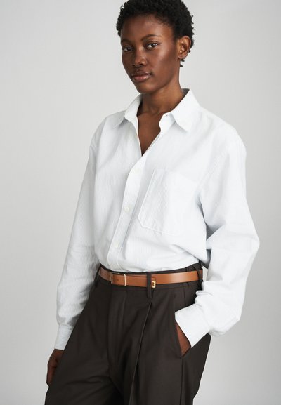 Λευκό πουκάμισο με ρίγες και ένα μόνο τσέπη, σε συνδυασμό με σκοτεινές πλισέ παντελόνες και μια καφέ ζώνη με χρυσή πόρπη.