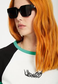 Schwarze übergroße quadratische Sonnenbrillen mit glänzender Oberfläche. Ein weißes und schwarzes Shirt mit grünen Akzenten und einem schwarzen Designdetail auf der Brust.