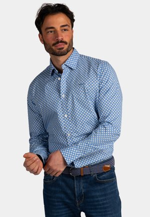 Man met donker haar en baard, gekleed in een blauwe blouse met lange mouwen en witte stippen, donkere spijkerbroek en een riem met patroon.