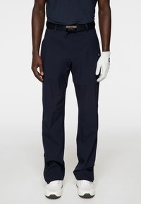 Marinblå golfbyxor i lätt material, med dragkedja vid ankeln, ett texturerat svart bälte, och tillsammans med vita golfskor och handskar.
