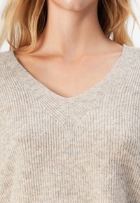 Mujer vestida con un suéter de punto acanalado beige con escote en V, que muestra el cuello, las clavículas y parte del cabello rubio.