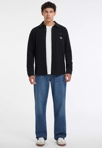 Giovane uomo in piedi, che indossa una camicia nera con bottoni sopra una maglietta bianca, jeans blu larghi e sneaker bianche con suola in gomma.