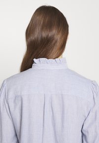 Personne aux longs cheveux bruns vue de dos portant une chemise fine rayée bleu clair et blanc avec un col à volants.