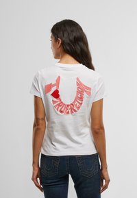 Witte katoenen t-shirt met daarop rode geborduurde tekst "TRUE RELIGION" en een hartontwerp op de achterkant. Korte mouwen en een ronde hals.