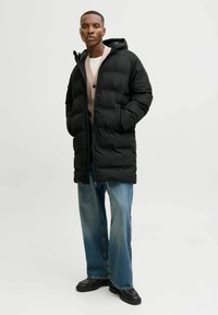Schwarze Daunenjacke mit Kapuze, ausgestattet mit einem Reißverschluss und strukturiertem, isoliertem Design. Trägt man über einem beigen Cardigan und weit geschnittenen Jeans.